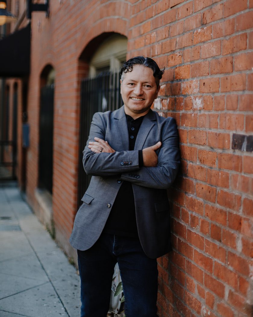 Mario Seijo, arms folded, leaning against a brick wall on a Seattle street.