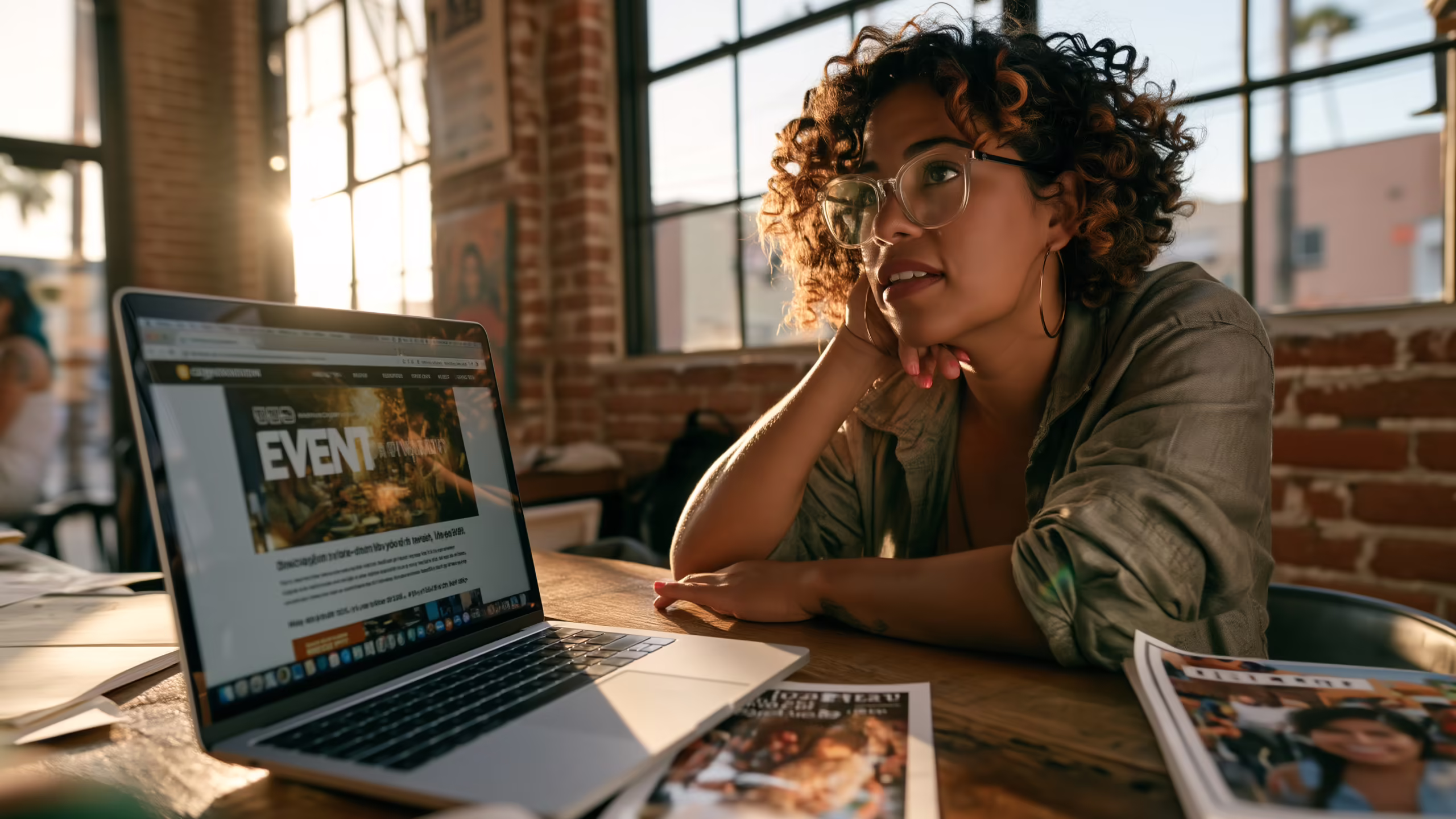 A nonprofit director rests her chin in her hand beside a laptop showing her organization's homepage, still promoting a past event, with printed event flyers on the table.